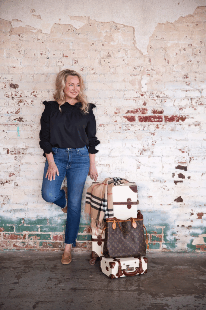 International Travel Agent Greta Meschke of Greta & Co. Travel Design standing beside luxury luggage in front of a rustic brick wall, representing global travel style and expertise.